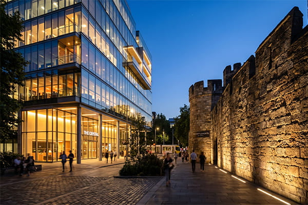 A modern glass building illuminated with wall washing and a historic stone wall lit with wall grazing